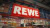 Großes rotes REWE-Logo hängt über Regalen im Lagerbereich.